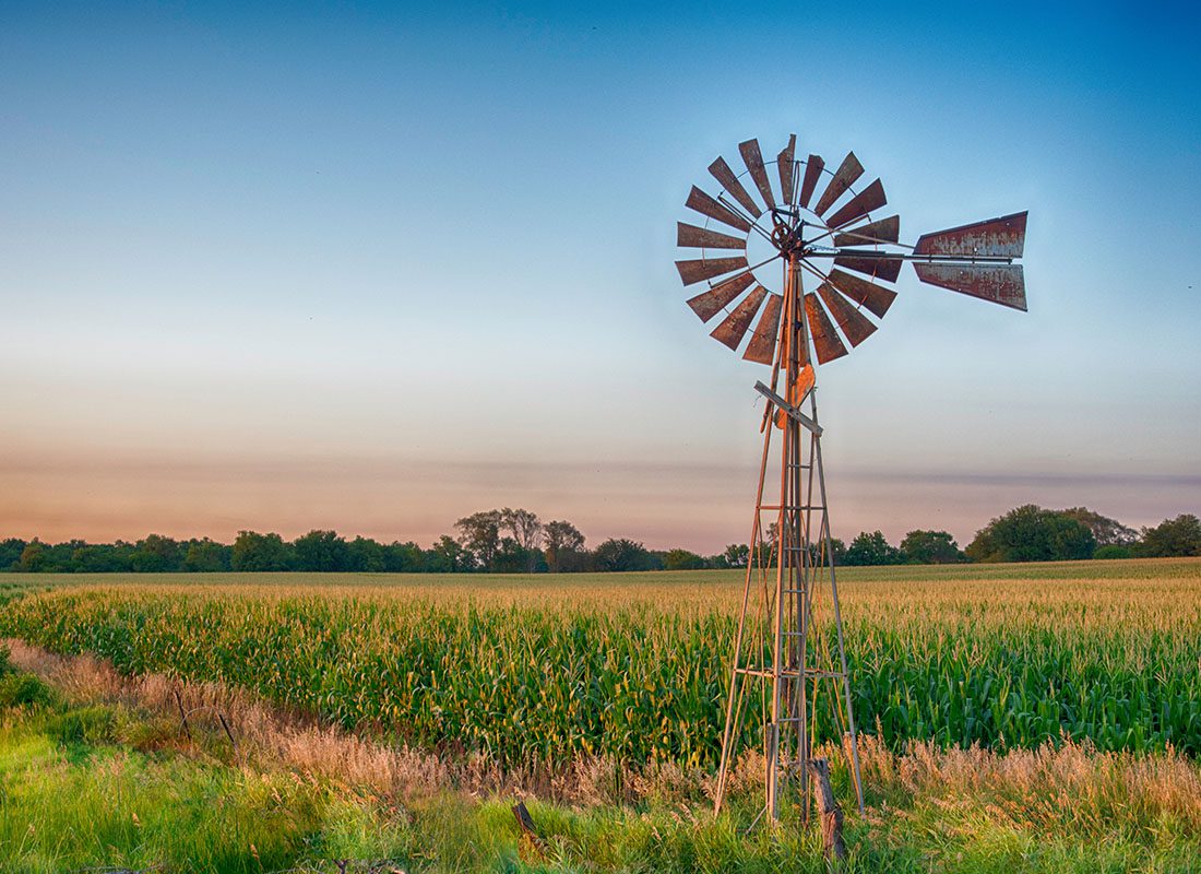 About Our Agency - Closeup View of an Antique Style Winmill on a Farm with Corn Growing in the Background at Sunset with a Clear Blue Sky