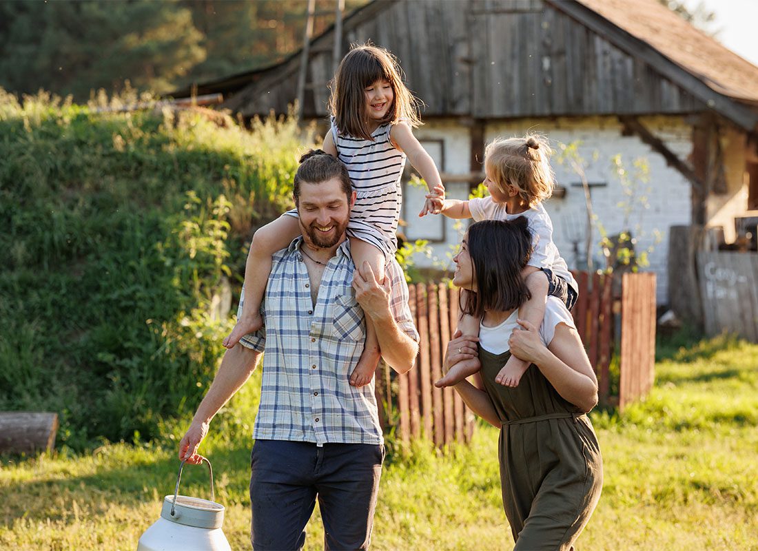 Personal Insurance - Portrait of Cheerful Young Parents Giving Their Two Daughters Piggyback Rides as They Walk Through Their Rural Property on a Sunny Day